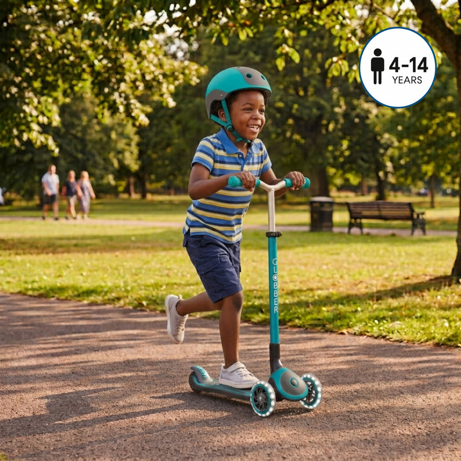 A young boy riding a teal and white scooter on a paved path in a sunny park, wearing a teal helmet. A white circular badge in the top right corner displays a person icon and the text '4-14 YEARS'.